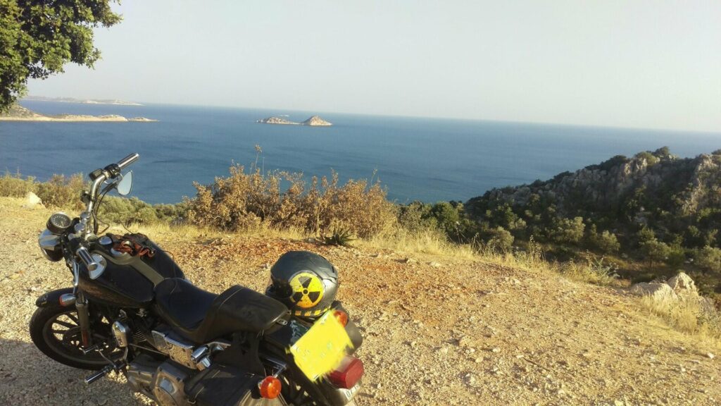 A motorcycle sits perched on the roadside in coastal Turkiye with the sea and islands in the back ground.